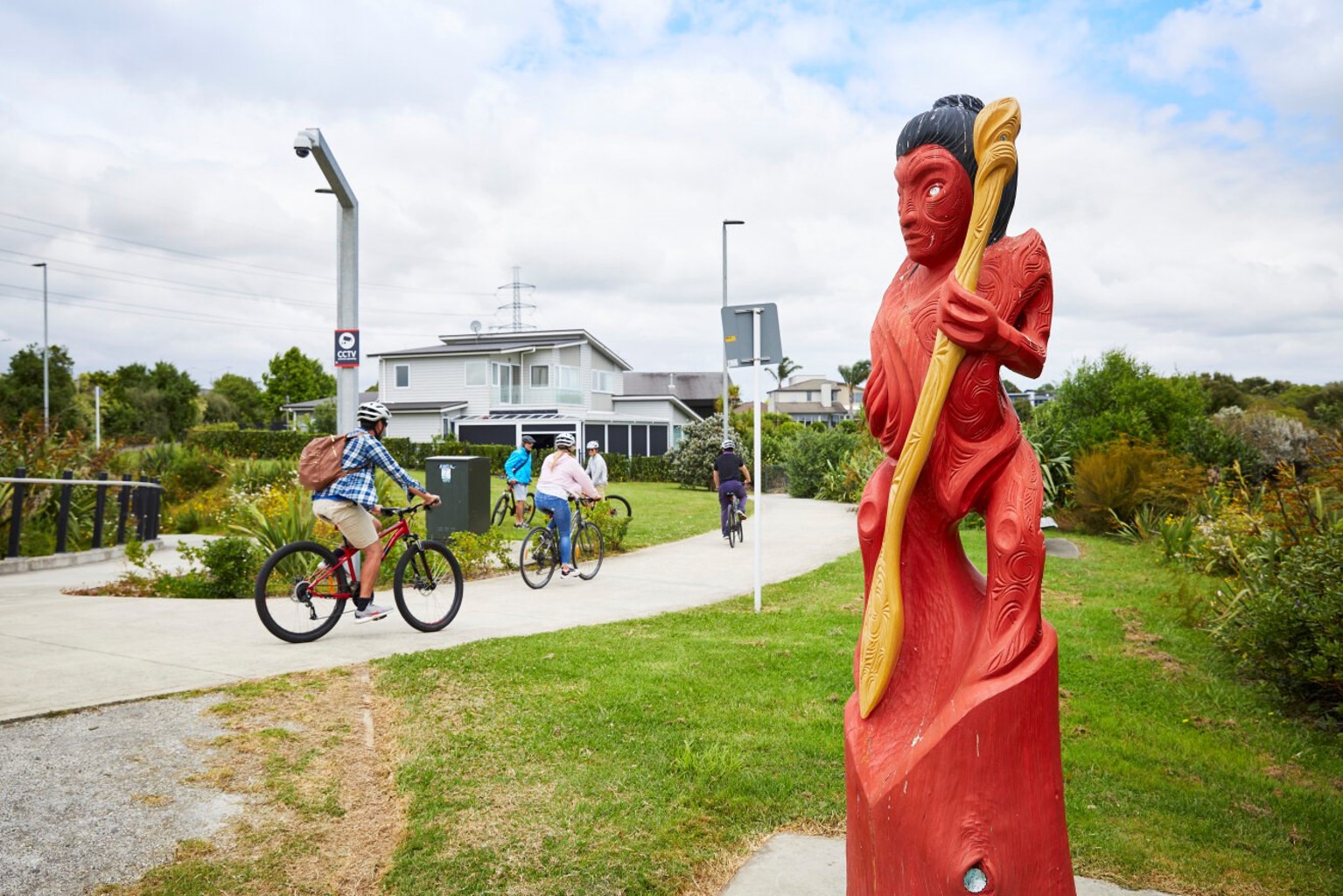 Cyclists riding along a concrete path with a Māori statue in the foreground