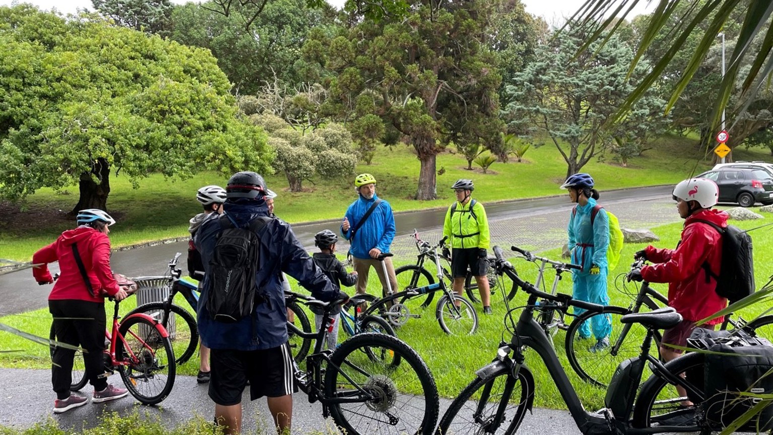 A group of bikers standing in an outdoor area with a bike track, listening to instructions on a guided bike walk. 