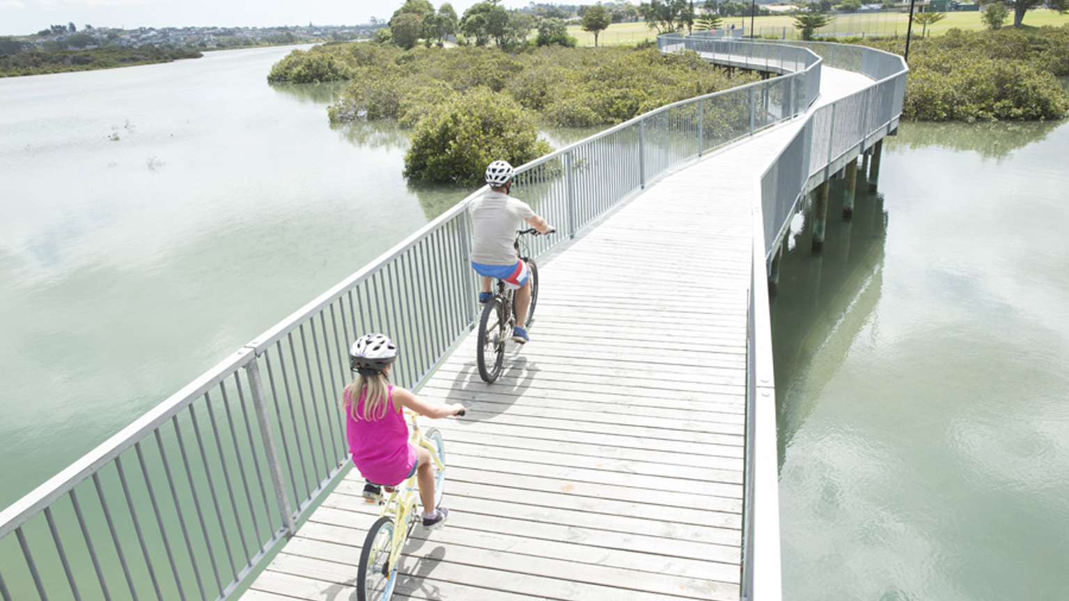 A father and daughter ride bikes across a bridge over calm, green water to a nearby park.