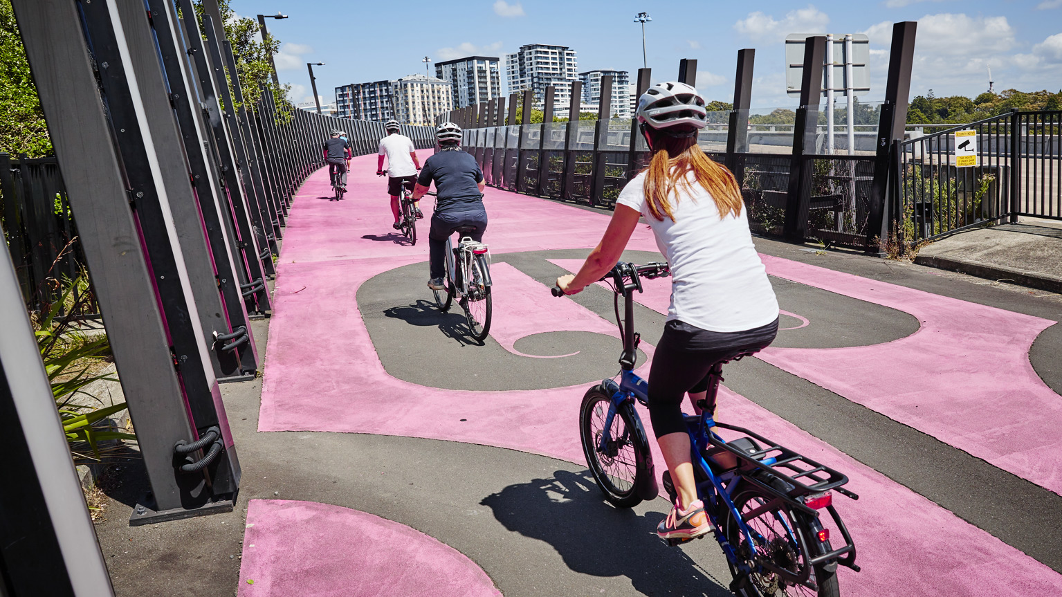 A woman on a bike follows a line of people on bikes ahead of her. They a cycling along Auckland's pink Te Ara I Whiti or Lightpath.