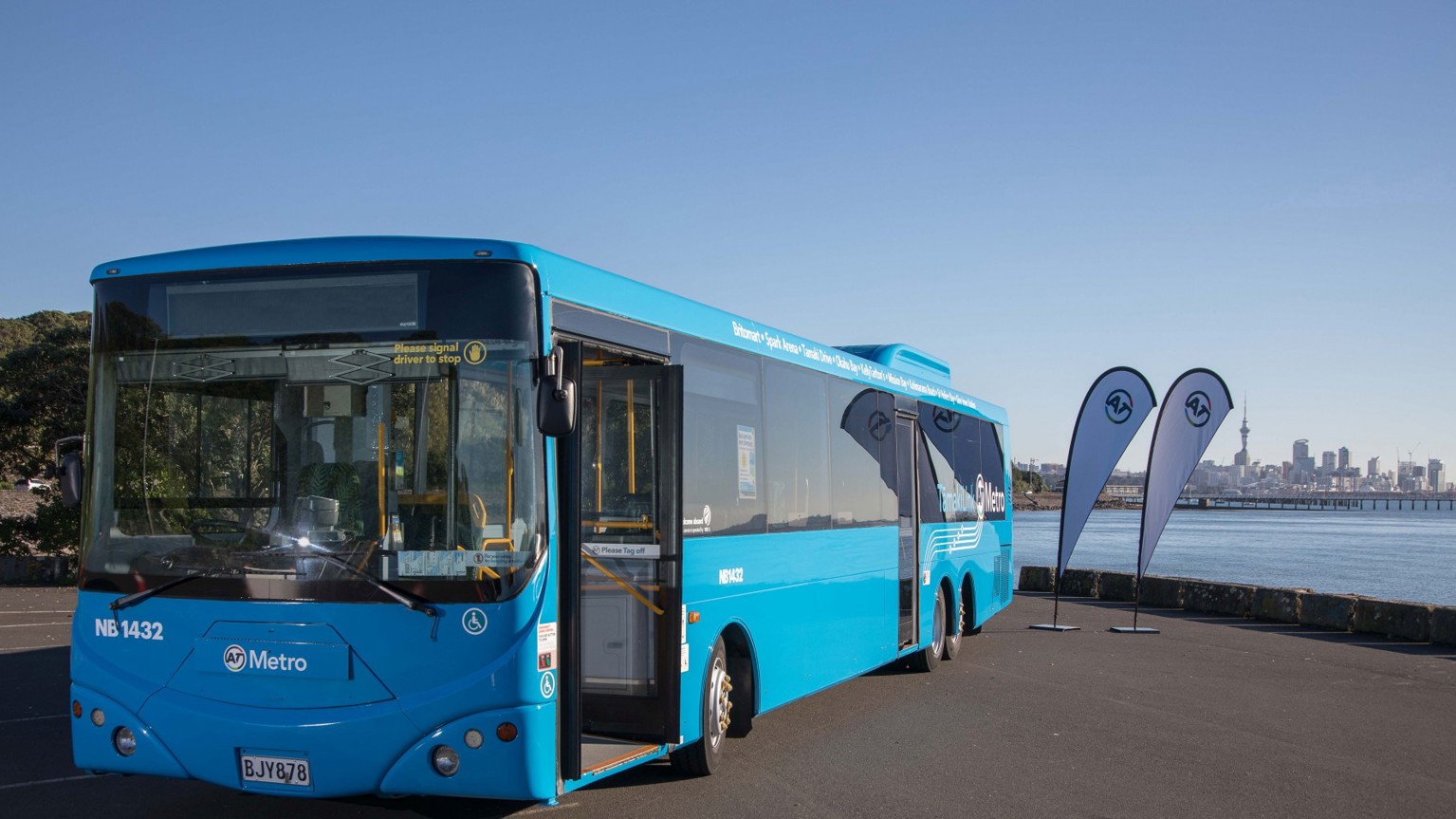 Tāmaki Link blue bus parked on the side of the road with the Auckland skyline in the background.