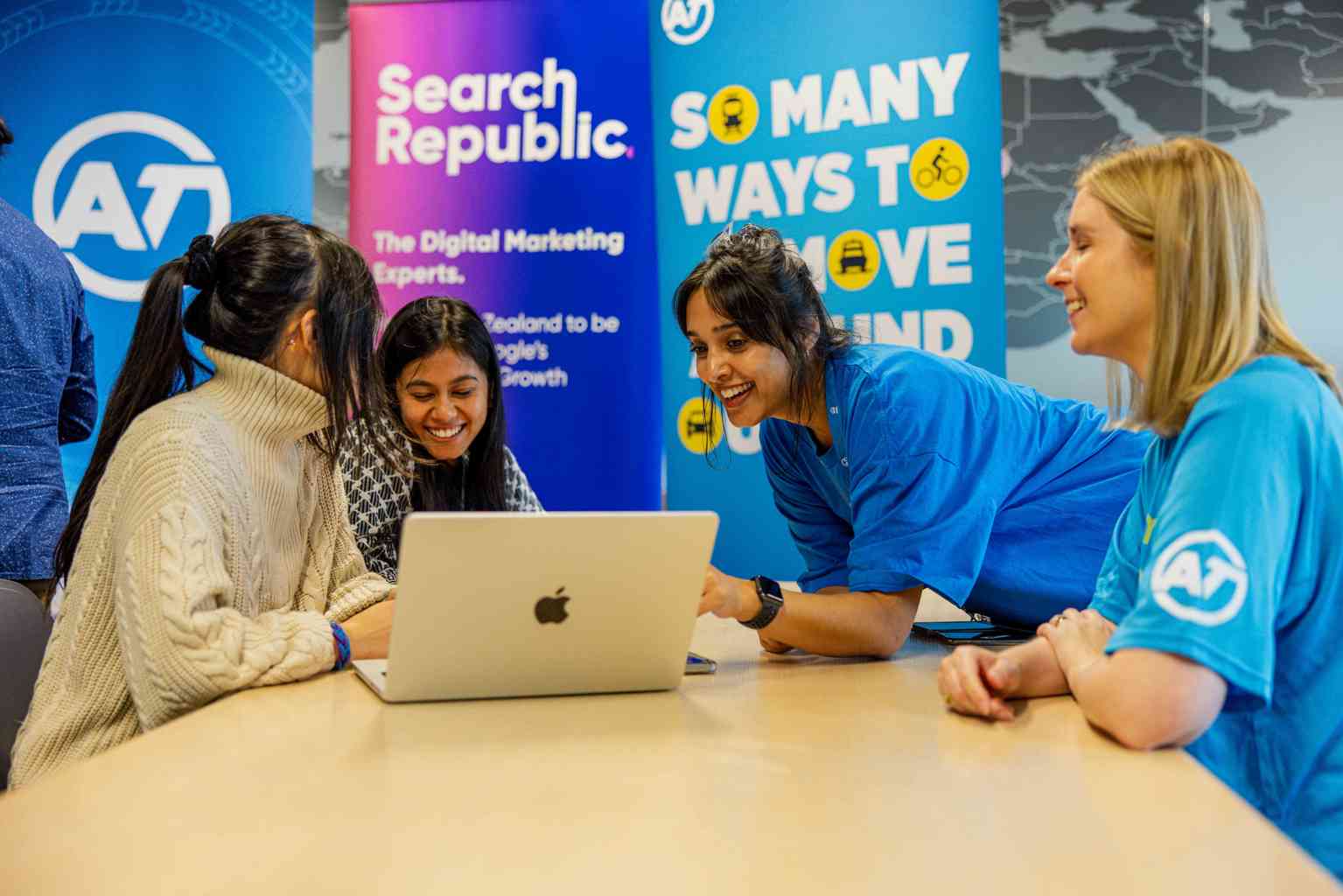 Four women gathered around an Apple laptop, one leaning over and pointing at the screen. Two women in AT logo shirts are helping the other 2, and branded banners hang in the background.